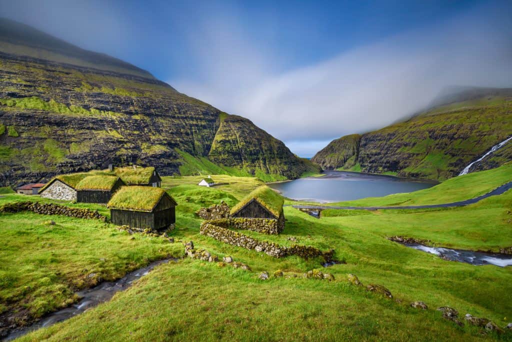 Grass-roofed cottages on a green hillside with a stone wall, adjacent stream, and distant lake, surrounded by mountains under a cloudy sky.