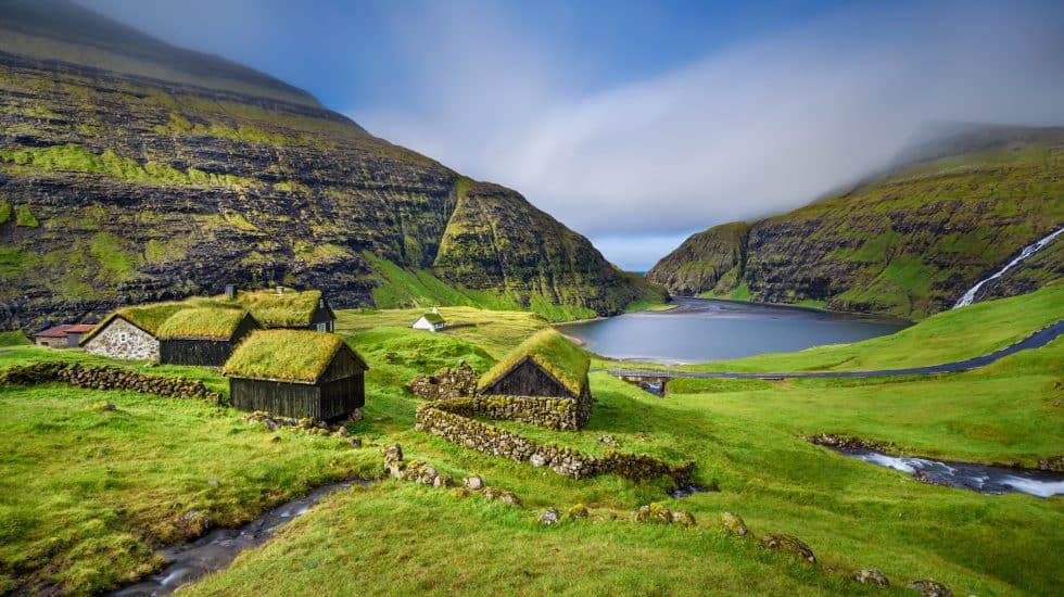 Remote Icelandic village with traditional grass-roofed cottages by a pristine lake surrounded by steep green cliffs and misty mountains.