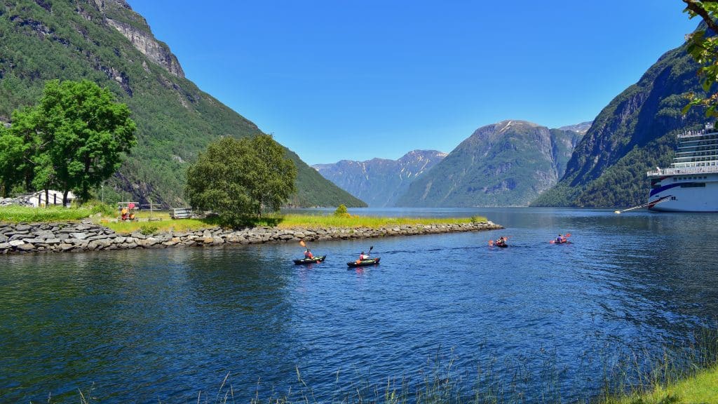 Kayakers paddle on a calm blue fjord surrounded by steep green mountains under a clear blue sky, with a large cruise ship docked nearby.