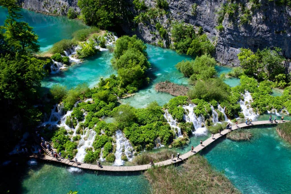 People walk on a wooden boardwalk winding through vibrant turquoise lakes and cascading waterfalls surrounded by lush greenery in a scenic landscape.