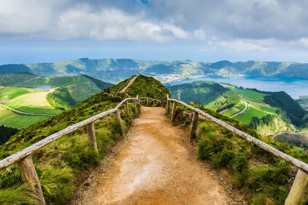 A dirt path with wooden railing leads through lush green hills overlooking a lake and distant mountains under a cloudy sky.
