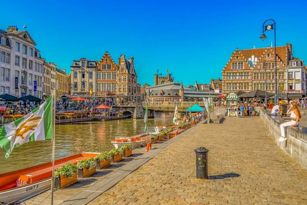 Canal-side view of historic buildings in Ghent, Belgium, with boats and outdoor cafes, sunny weather, and vibrant city atmosphere.