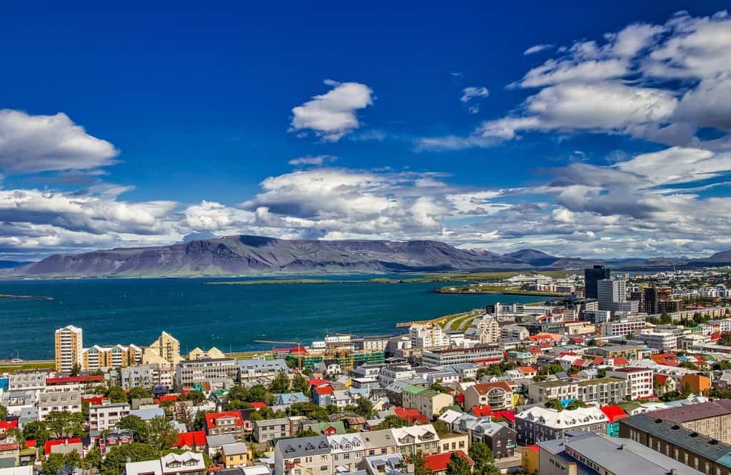 Vibrant cityscape of Reykjavik, Iceland, with colourful rooftops, modern buildings, and stunning Mount Esja in the background under a bright blue sky with scattered clouds.