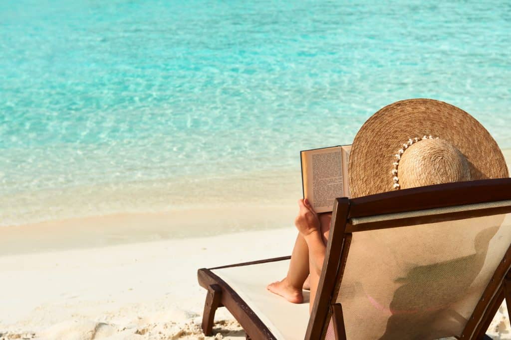 Person wearing a large straw hat reads a book while reclining on a beach chair facing the clear turquoise sea.