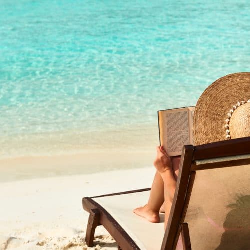 Relaxing beach scene with woman reading a book on a lounge chair under a straw hat by turquoise sea, perfect for travel relaxation and holiday escape.