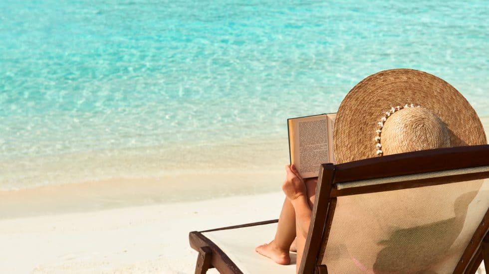 Relaxing beach scene with woman reading a book on a lounge chair under a straw hat by turquoise sea, perfect for travel relaxation and holiday escape.