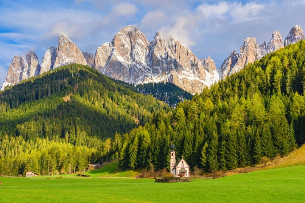 A small church stands in a green field surrounded by dense pine forests with the jagged, snow-tipped Dolomite mountains in the background under a partly cloudy sky.