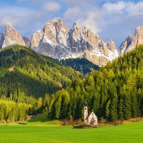 Vibrant mountain landscape with lush green forests, towering snow-capped peaks, and a charming white chapel nestled in a grassy meadow, showcasing the stunning scenery of the Dolomites in Italy.