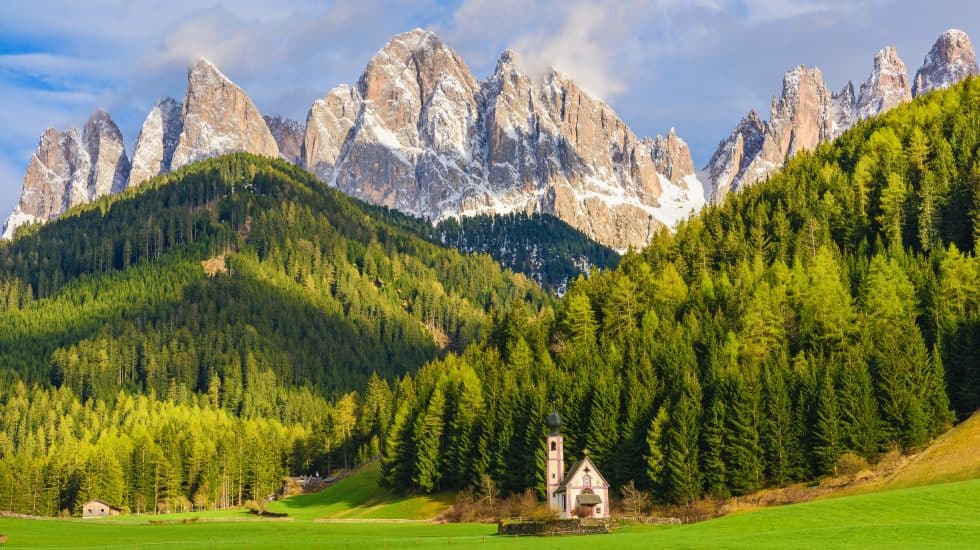 Vibrant mountain landscape with lush green forests, towering snow-capped peaks, and a charming white chapel nestled in a grassy meadow, showcasing the stunning scenery of the Dolomites in Italy.