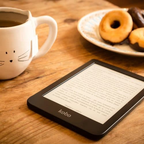 Cozy coffee mug with a cat face design, a plate of assorted donuts, and an e-reader on a wooden table, perfect for relaxing travel reading and enjoying sweet treats during a holiday break.