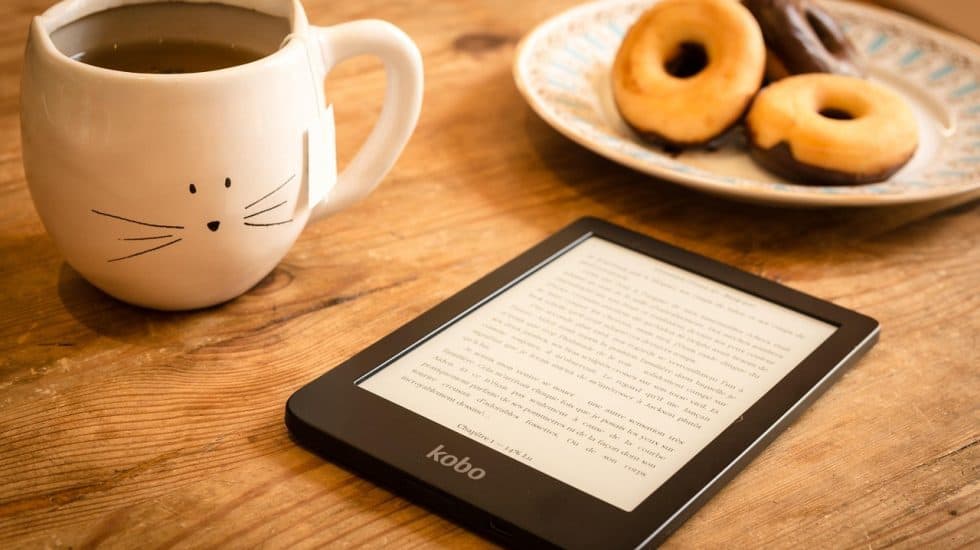 Cozy coffee mug with a cat face design, a plate of assorted donuts, and an e-reader on a wooden table, perfect for relaxing travel reading and enjoying sweet treats during a holiday break.