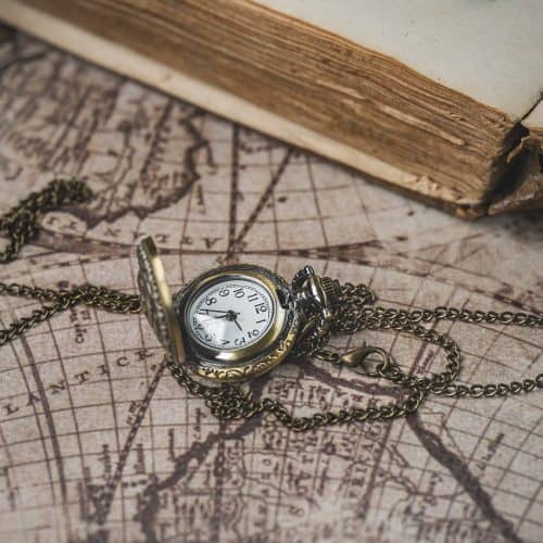A vintage pocket watch with a chain is open on an old world map, near the edge of an aged, partially opened book.