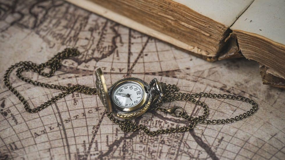 A vintage pocket watch with a chain is open on an old world map, near the edge of an aged, partially opened book.