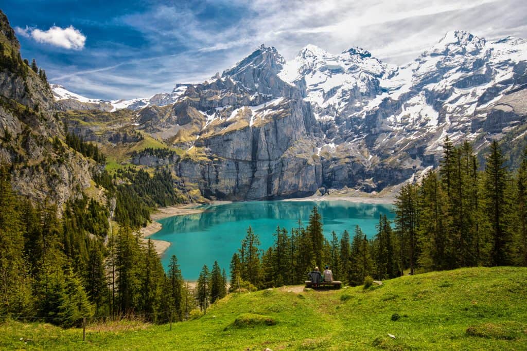Two people sit on a bench overlooking a turquoise alpine lake surrounded by snow-capped mountains and lush pine trees under a bright blue sky.