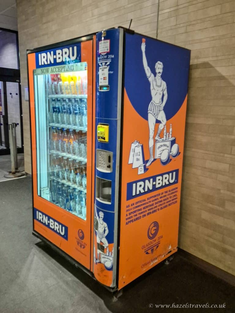 IRN-BRU vending machine at Glasgow 2014 Commonwealth Games, featuring vintage-style mascot design and bright orange branding, located indoors for quick refreshment access.