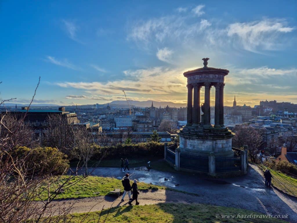 View from Calton Hill in Edinburgh at sunset, showing the Dugald Stewart Monument, city buildings, people walking, and a bright sky with scattered clouds.