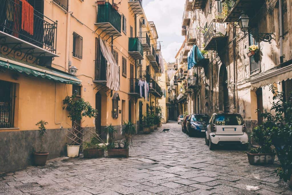 Narrow cobblestone street lined with parked cars, potted plants, laundry hanging on balconies, and yellow buildings in daylight.