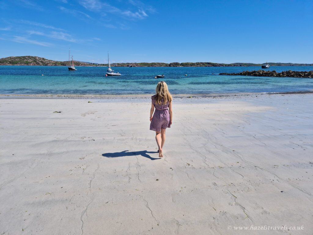 A woman in a dress walks barefoot on a sandy beach toward the sea, with sailboats anchored in the water under a clear blue sky.