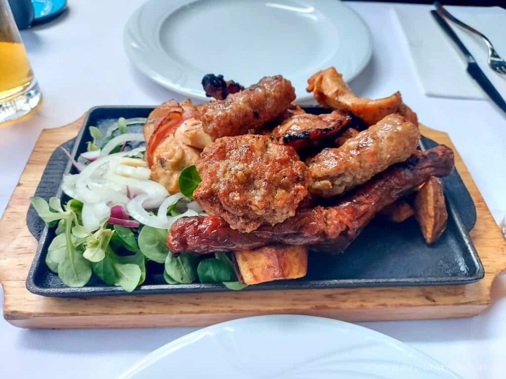 A platter of traditional Serbian grilled meats including ćevapi and sausages, served on a wooden tray with sliced onions, greens, and roasted potatoes, set on a white tablecloth next to an empty plate and cutlery.