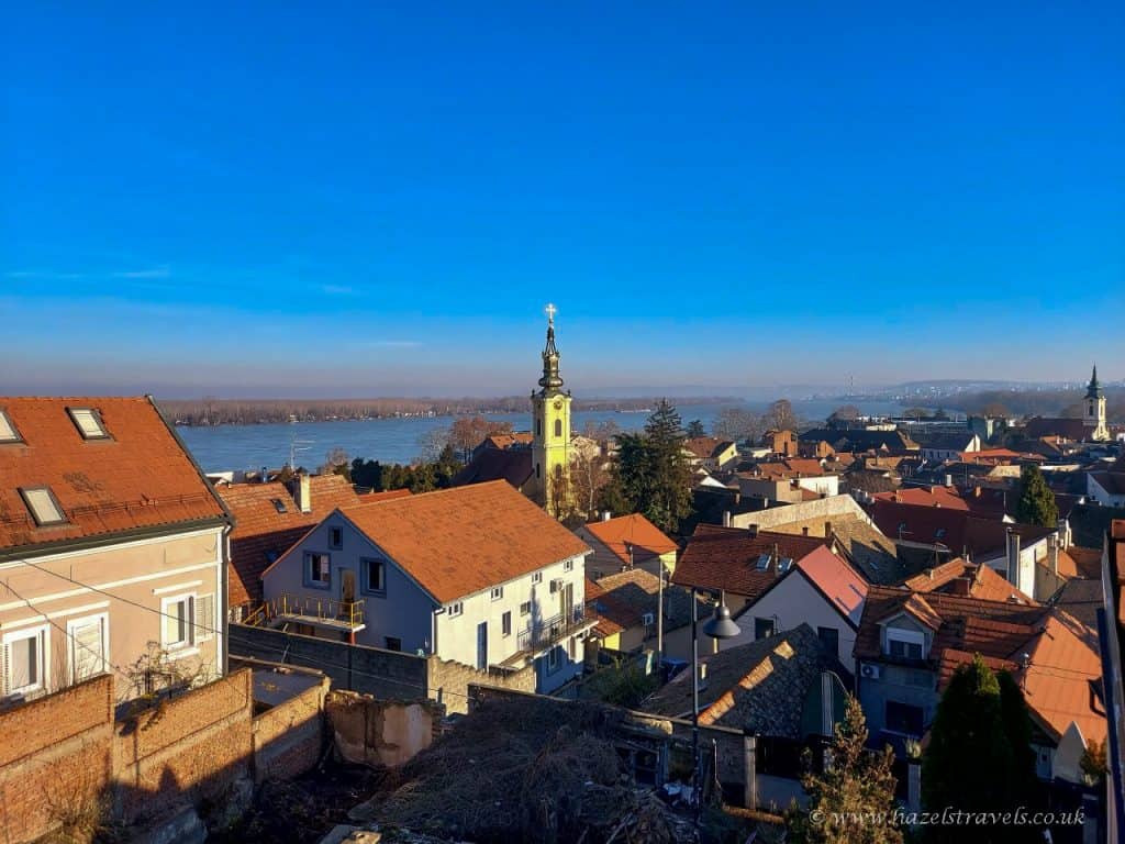 Scenic view over the rooftops of Zemun, Belgrade, with a yellow church tower in the foreground and the Danube River glistening under a clear blue sky in the background.