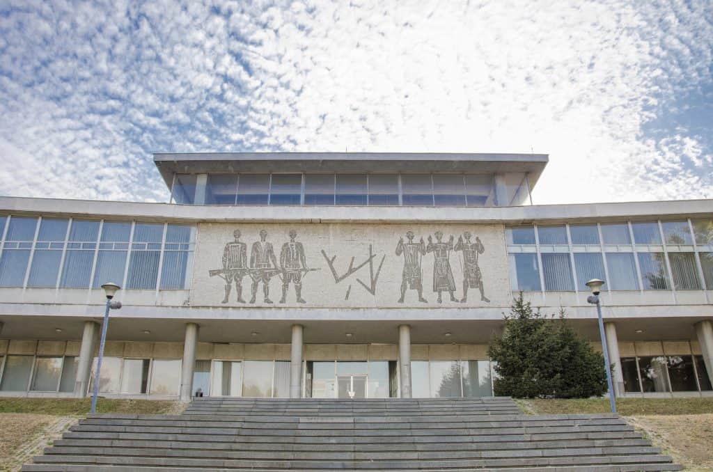 Front view of the Museum of Yugoslavia in Belgrade, with a wide staircase leading up to a modernist white building featuring tall windows and a central mural of stylized human figures under a partly cloudy sky.