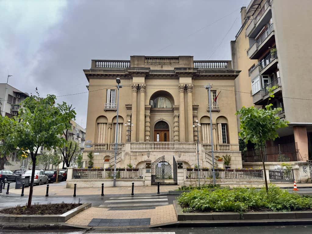 Exterior view of the Nikola Tesla Museum in Belgrade, a cream-colored neoclassical building with tall arched windows, decorative columns, and a gated entrance, set between modern apartment buildings on a cloudy day.