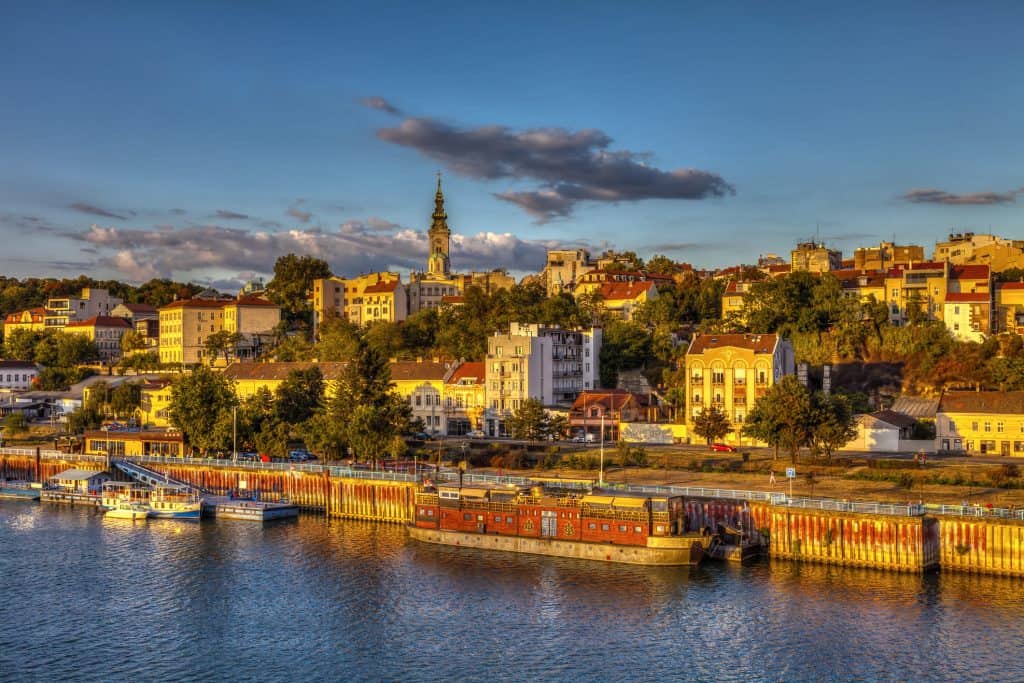 Golden hour view of Belgrade's historic waterfront, showing colorful buildings on a hillside, with the spire of St. Michael's Cathedral rising above. Houseboats and splavs (floating bars) line the Danube River under a blue sky with scattered clouds.