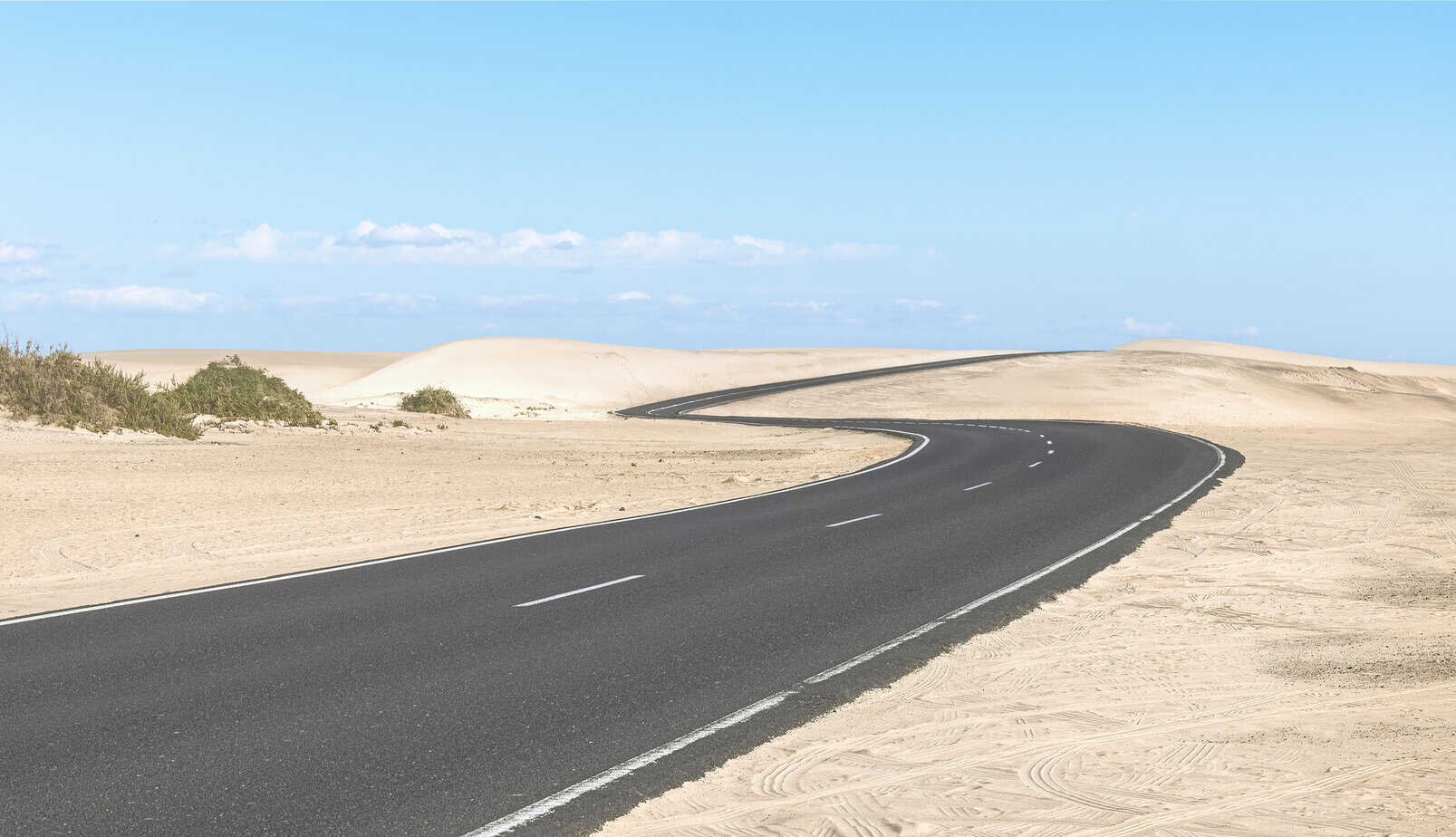 A paved road curves through a sandy desert landscape under a clear blue sky, with small shrubs visible on the left side.
