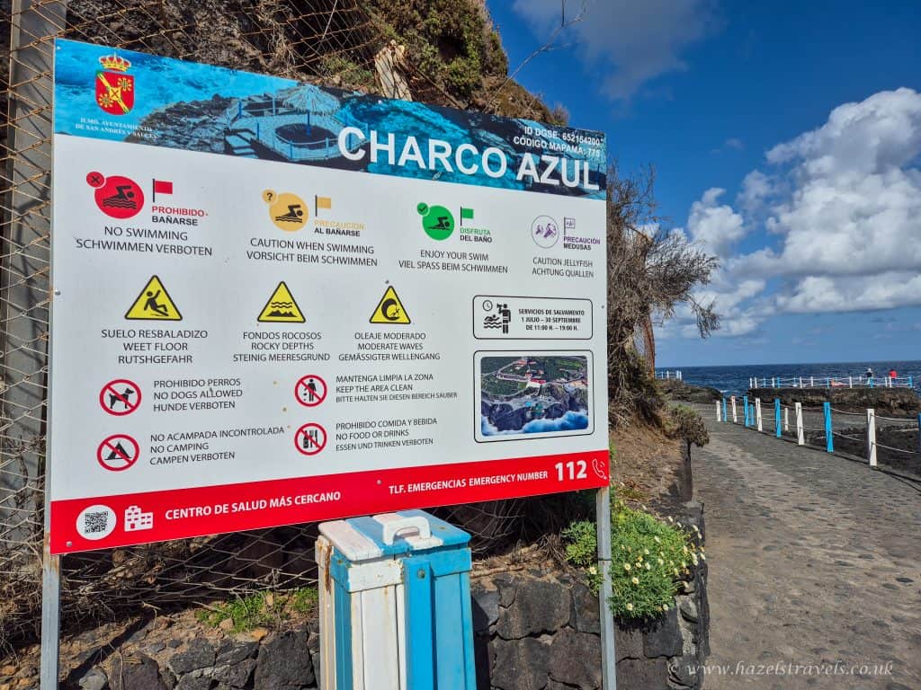 No swimming prohibited sign on blue and white information board at Charco Azul, Tenerife, Canary Islands, Spain, with ocean view and coastal pathway in the background.