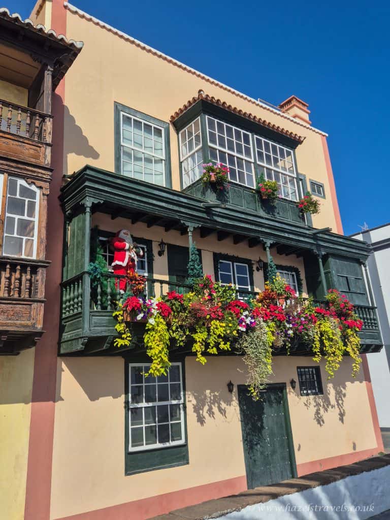 Bright colourful building with a green painted balcony and festive decorations, including a Santa figure and vibrant flower arrangements, under a clear blue sky in a scenic travel destination.