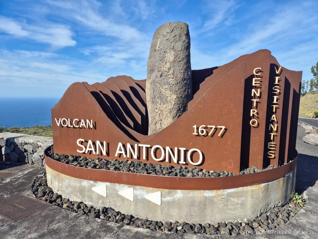 Rustic metal sign for Volcán San Antonio Visitor Center, featuring a central stone and the year 1677, with ocean and sky visible in the background.