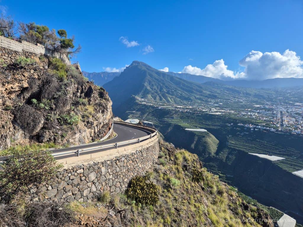 A winding mountain road curves along a steep rocky cliff, overlooking a green valley with distant mountains under a clear blue sky.