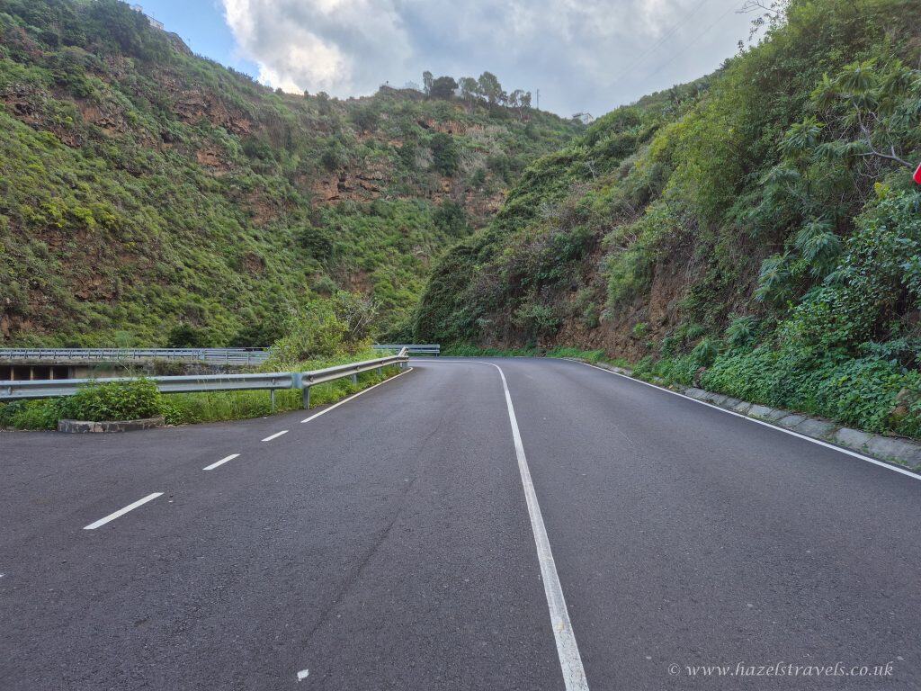 A paved road curves through green, hilly terrain with a metal guardrail on the left and dense vegetation on both sides.
