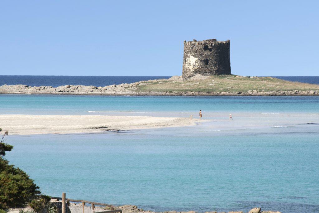 Serene coastal scene with an ancient stone tower on a small island, clear turquoise waters, sandy beach, and people enjoying the seaside in Cornwall, UK.