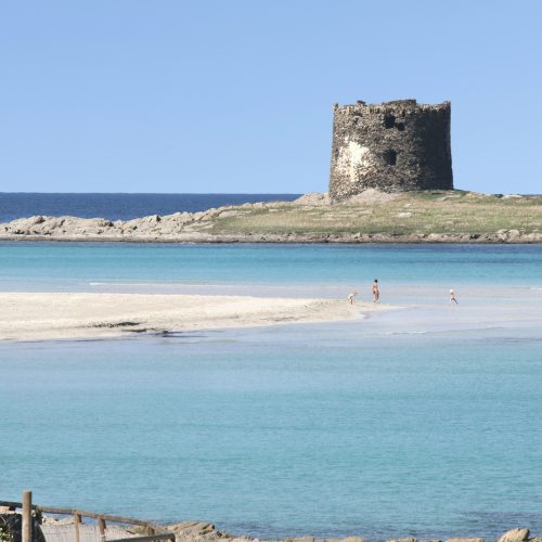 Serene coastal scene with an ancient stone tower on a small island, clear turquoise waters, sandy beach, and people enjoying the seaside in Cornwall, UK.