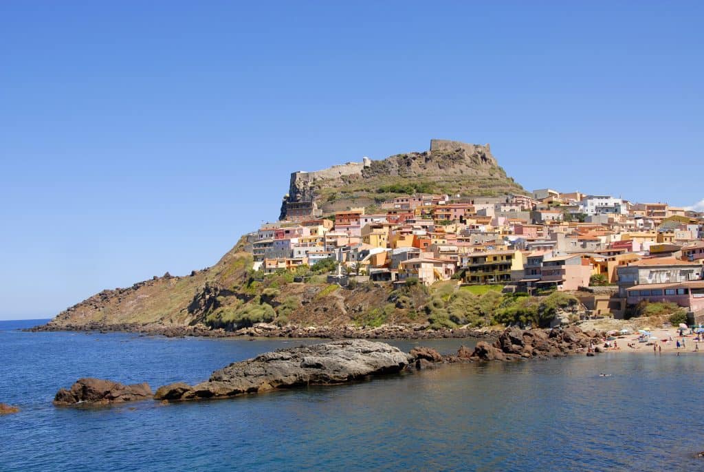 Colourful coastal village on a hillside with a historic fortress at the top, overlooking the sea and beach on a clear sunny day, perfect for travel and holiday photography.