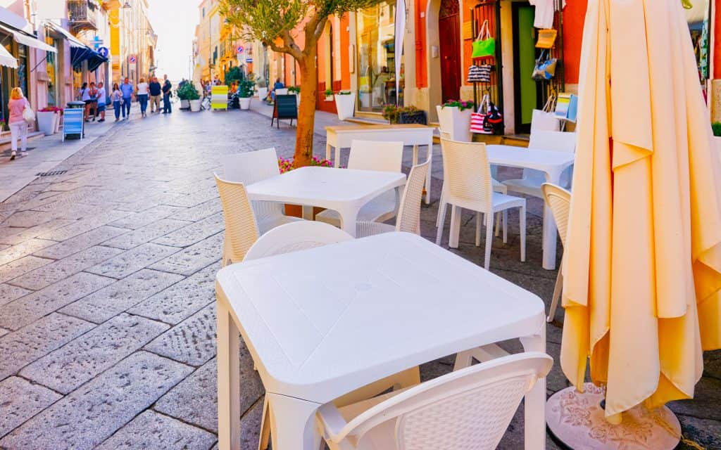 Outdoor café tables and chairs line a cobblestone street with shops on both sides; a few people are walking in the distance.