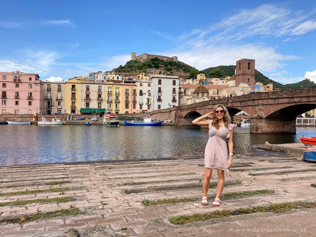 Colourful waterfront in Italy with historic buildings, a stone bridge, and a hilltop castle, featuring a woman enjoying a day by the canal. Ideal for travel and holiday photography.