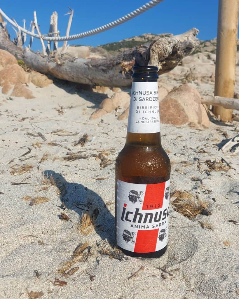 Bottle of Ichnusa beer on Sardinian beach with driftwood and rocks in the background.