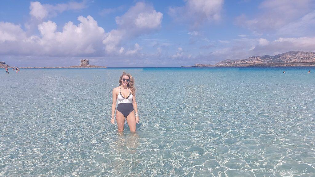 A woman in a swimsuit stands in shallow, clear blue water at a beach with distant islands and a tower visible under a partly cloudy sky.