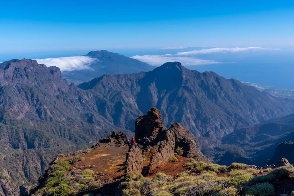 Mountain landscape with rugged peaks, green vegetation in the foreground, and clouds covering distant valleys under a clear blue sky.