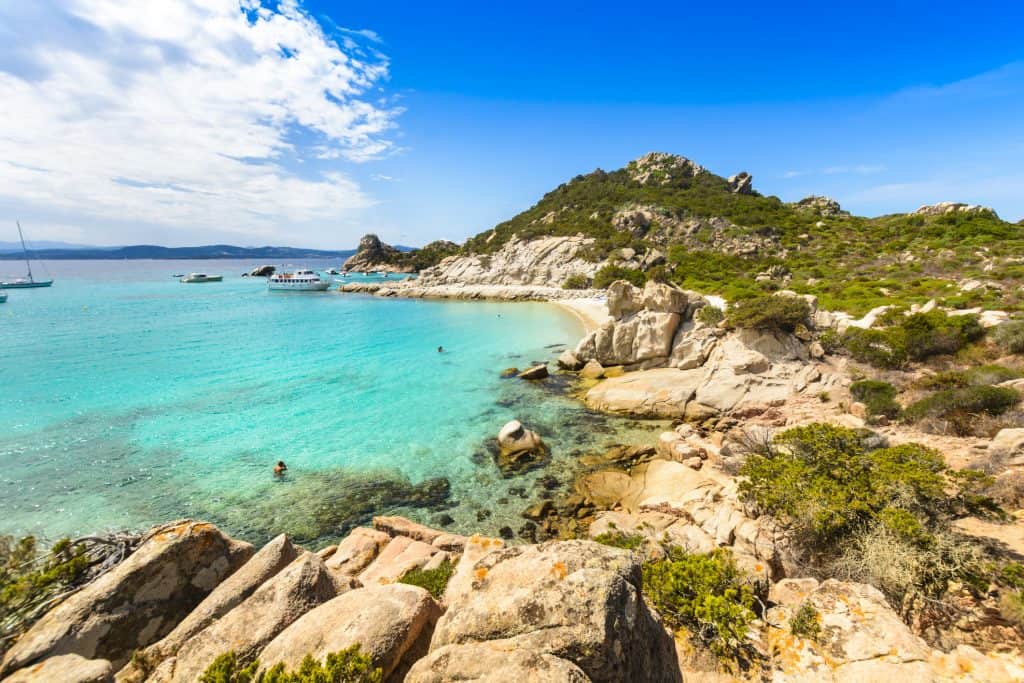 Sunny Mediterranean beach with turquoise waters, rocky shoreline, lush green hills, and boats anchored near the coast, perfect for scenic coastal holidays and relaxing seaside escapes.