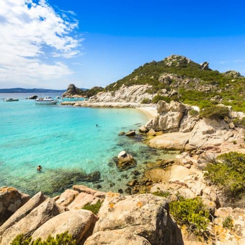 Sunny Mediterranean beach with turquoise waters, rocky shoreline, lush green hills, and boats anchored near the coast, perfect for scenic coastal holidays and relaxing seaside escapes.