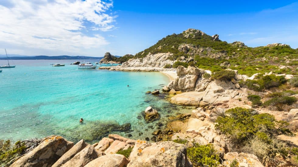 Sunny Mediterranean beach with turquoise waters, rocky shoreline, lush green hills, and boats anchored near the coast, perfect for scenic coastal holidays and relaxing seaside escapes.
