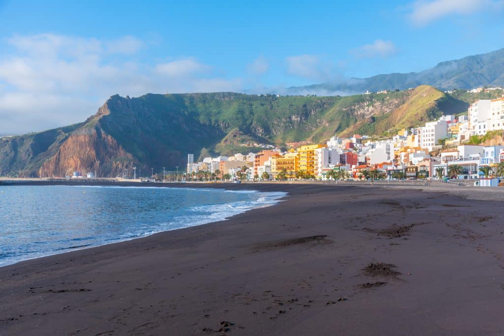 A sandy beach with dark volcanic sand stretches alongside a coastal town with colorful buildings, set against green hills and mountains under a partly cloudy sky.