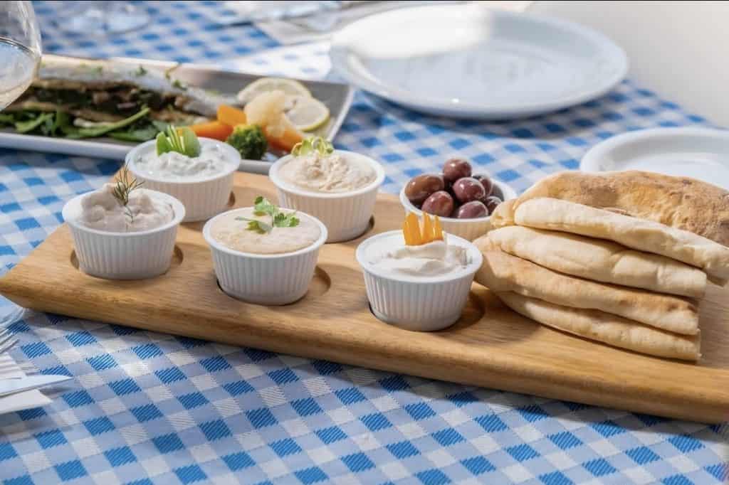 A wooden board with five small dishes of assorted dips, a pile of pita bread, and a few olives, set on a blue-checkered tablecloth in a restaurant in Paphos, Cyprus.