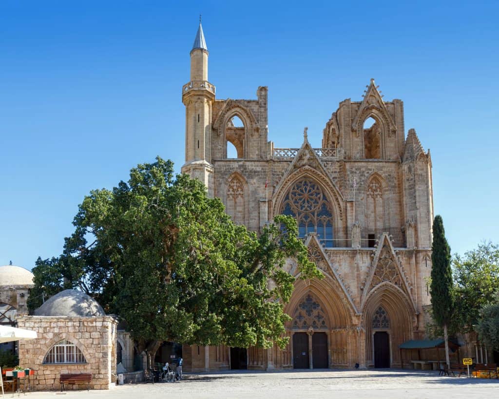 A large Gothic-style stone building in Famagusta, Cyprus with arched windows and a minaret on the left side, set against a clear blue sky and surrounded by trees.