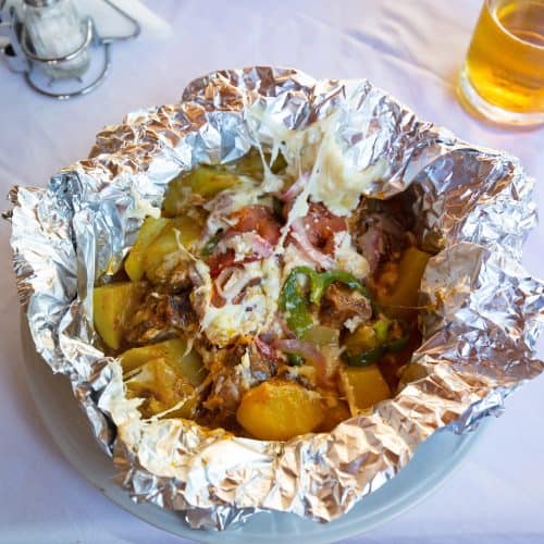 Crispy baked seafood casserole in foil with vegetables and cheese, served on a white table with beer in the background.