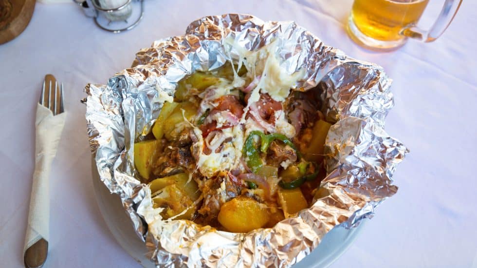 Crispy baked seafood casserole in foil with vegetables and cheese, served on a white table with beer in the background.