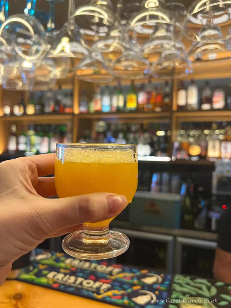 Refreshing yellow cocktail in a glass at a lively bar deep in focus with hanging wine glasses overhead and shelves of spirits in the background, perfect for travel and leisure content.